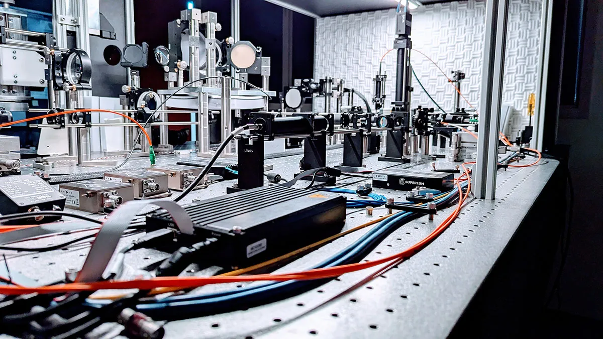 Close-up of an adaptive optics scanning laser ophthalmoscope (AOSLO) on an optical breadboard, showing mirrors, lens mounts, and fiber-coupled light sources.